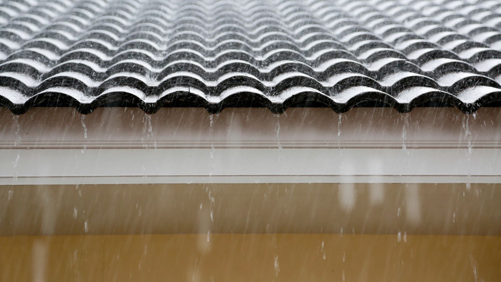 Lluvia sobre un tejado de tejas oscuras, con gotas de agua cayendo desde el borde sobre una franja blanca.